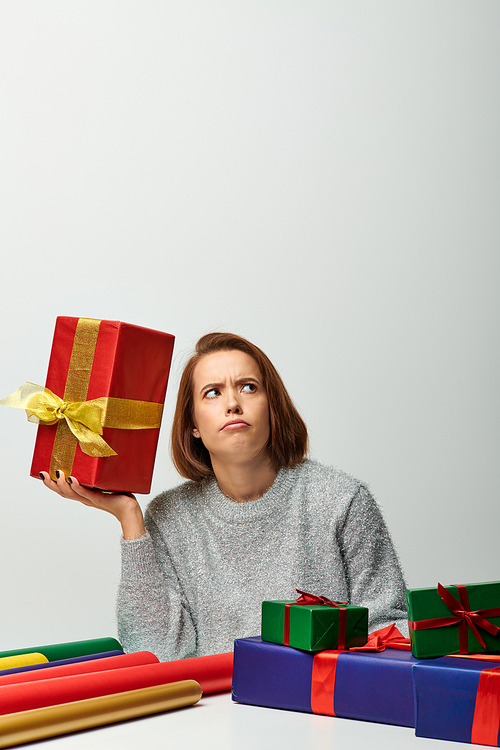 curious woman in knitted sweater holding Christmas present near colorful gift paper on grey backdrop