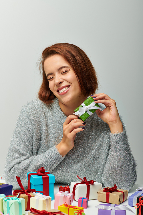 cheerful woman in cozy sweater holding tiny Christmas present near colorful wrapped gifts on grey