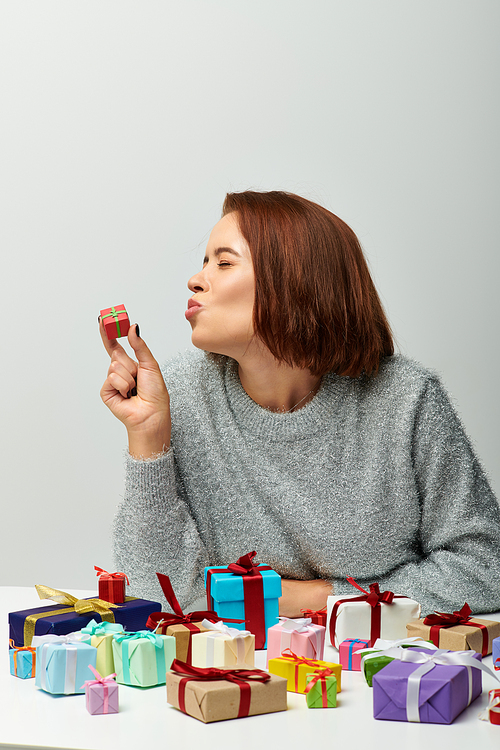 young woman in cozy sweater kissing tiny Christmas present near colorful wrapped gifts on grey