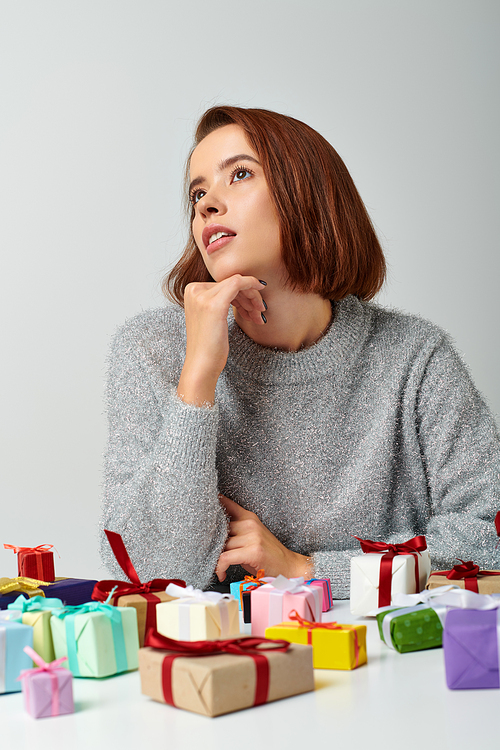pretty woman in winter sweater fantasizing among bunch of Christmas gifts on table, grey backdrop