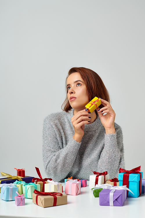 pretty woman in sweater fantasizing and holding Christmas gift near bunch of presents on table