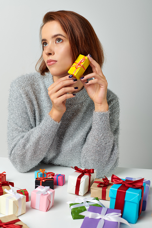 appealing woman in sweater fantasizing and holding Christmas gift near bunch of presents on table