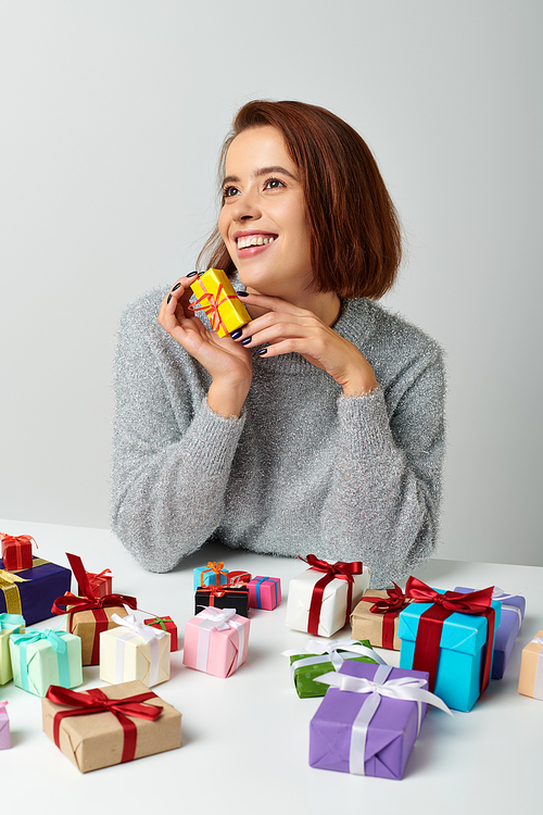 cheerful woman in sweater fantasizing and holding Christmas gift near bunch of presents on table