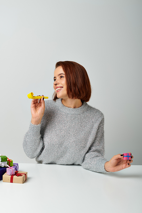 cheerful woman in sweater fantasizing and holding tiny Christmas gift and yellow party horn on grey