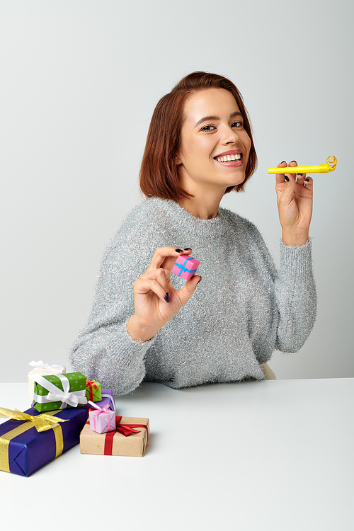 happy woman in sweater smiling and holding tiny Christmas gift and yellow party horn on grey