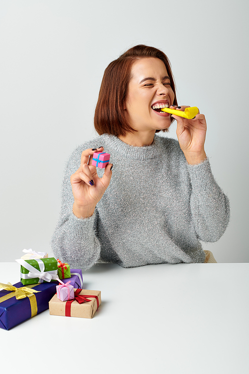 happy woman in sweater holding tiny Christmas gift and blowing yellow party horn on grey backdrop