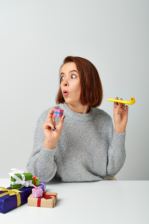 shocked woman in sweater holding tiny Christmas gift and yellow party horn on grey backdrop