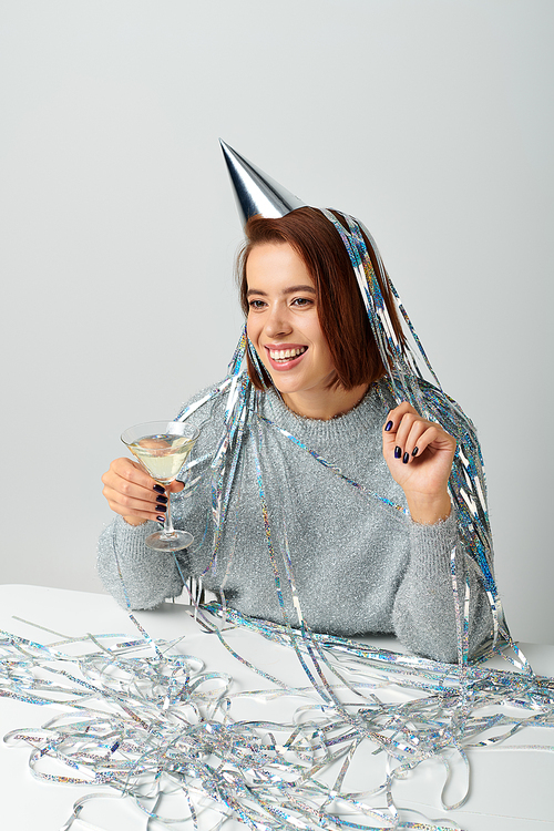 woman in party cap with silver tinsel on head holding glass of champagne on grey, Happy New Year
