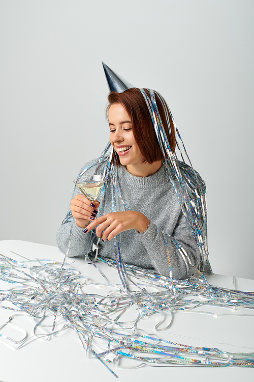 cheerful woman in party cap with tinsel on head holding glass of champagne on grey, Happy New Year