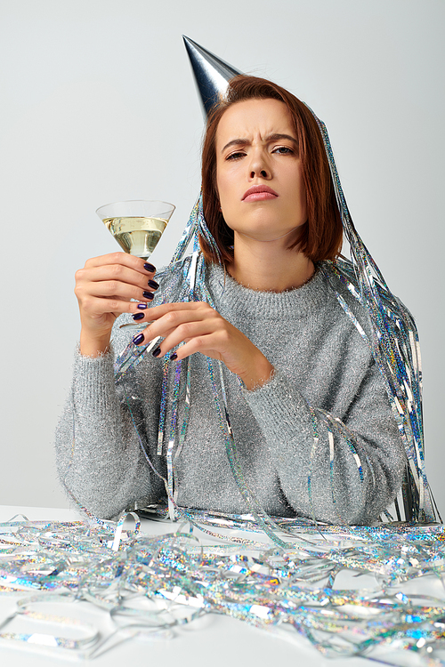 serious woman in party cap with tinsel on head holding glass of champagne on grey, New Year concept