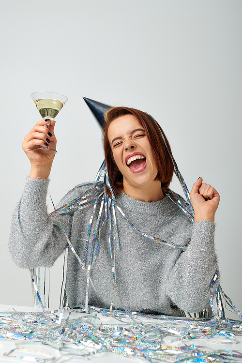 excited woman in party cap with tinsel on head holding glass of champagne on grey, Happy New Year
