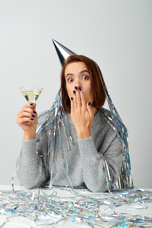 young woman in party cap with tinsel on head holding glass of champagne and covering face with hand