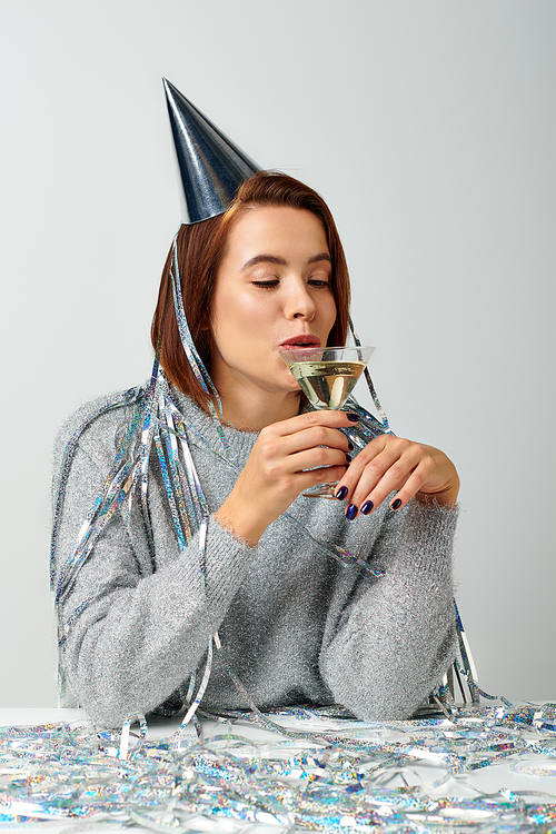 young woman in party cap with tinsel on head drinking champagne on grey backdrop, Happy New Year