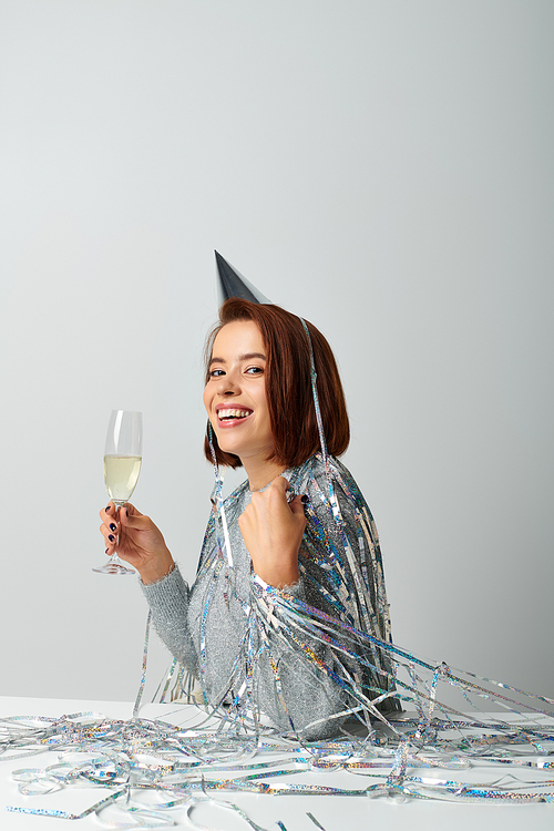joyful woman in party cap with tinsel on head holding glass of champagne while celebrating Christmas