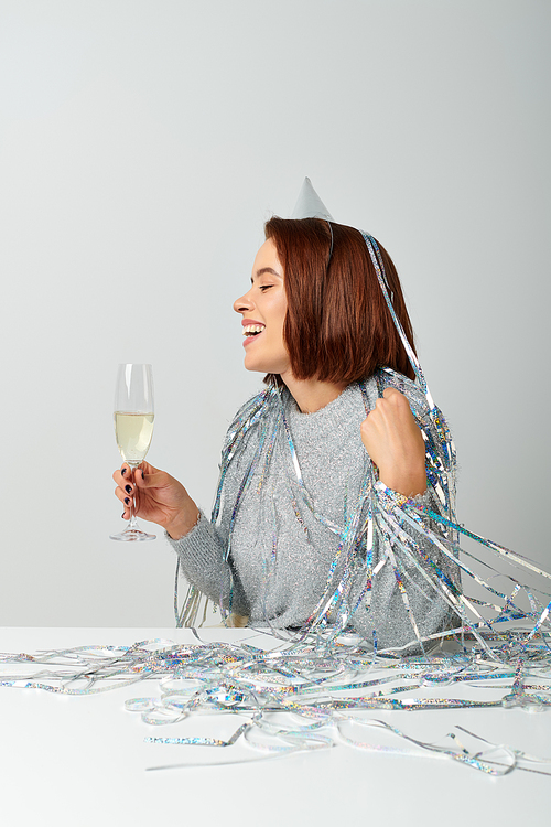 joyful woman in party cap and tinsel on head holding glass of champagne while celebrating New year
