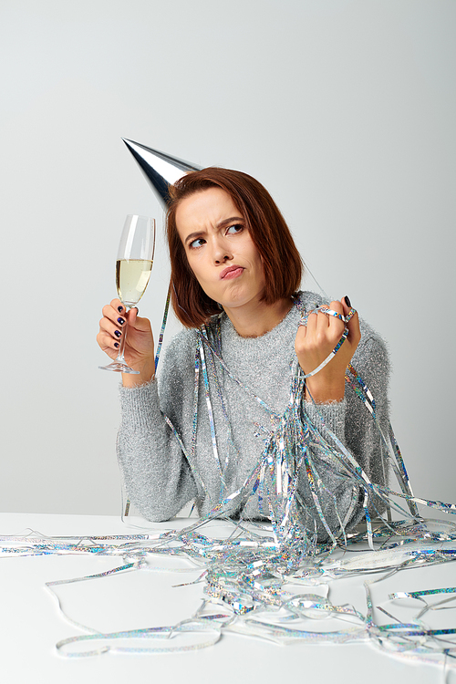 pensive woman in party cap and tinsel on head holding champagne glass while celebrating New year