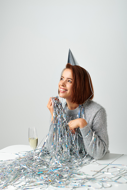 satisfied woman in party cap and tinsel smiling near champagne glass while celebrating New year