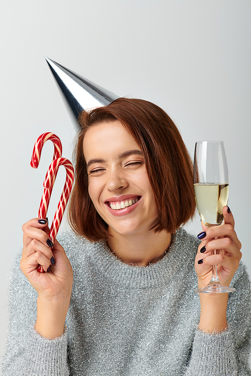 pleased woman in party cap holding champagne glass and candy canes on grey, Merry Christmas concept
