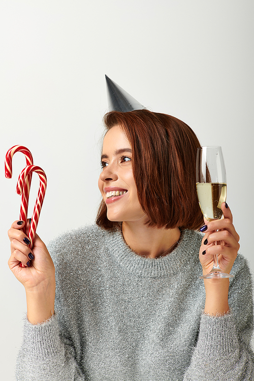 happy woman in party cap holding champagne glass and candy canes on grey, Merry Christmas concept