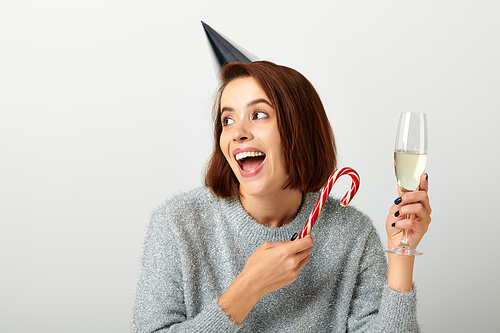 excited woman in party cap holding champagne glass and candy cane on grey, Merry Christmas