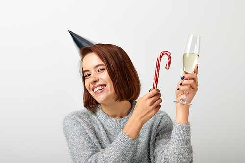 positive woman in party cap holding champagne glass and candy cane on grey, Merry Christmas
