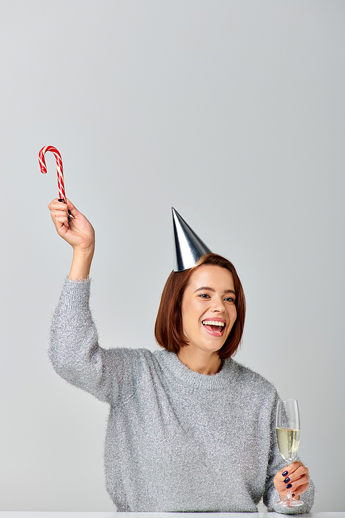 thrilled woman in party cap holding candy cane and champagne glass on grey backdrop, Merry Christmas