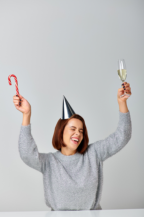 excited woman in party cap holding candy cane and champagne glass on grey backdrop, Happy New Year