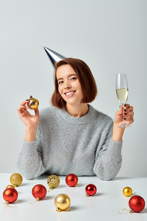 delighted woman in party cap holding champagne glass and bauble on grey, Merry Christmas concept