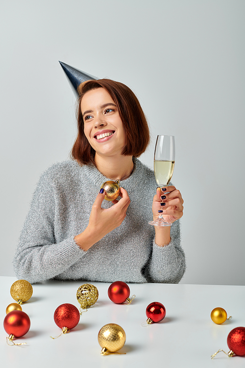pleased woman in party cap holding champagne glass and bauble on grey, Merry Christmas concept