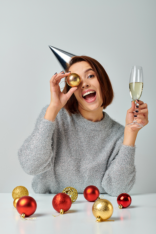 excited woman in party cap holding champagne glass and bauble on grey, Merry Christmas concept