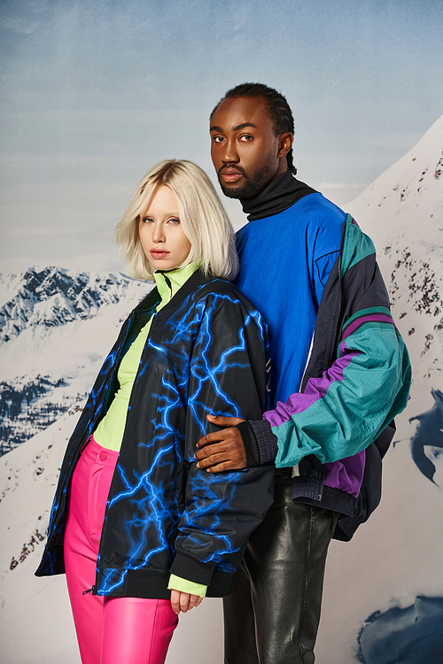 attractive young multicultural couple in stylish winter attire hugging and posing on snowy backdrop