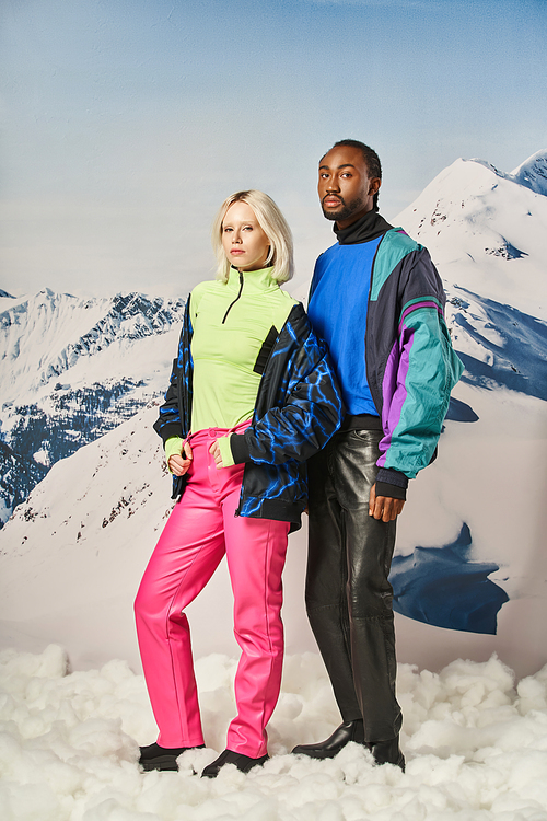 young stylish couple in vibrant winter outfits standing on snow with mountain on background