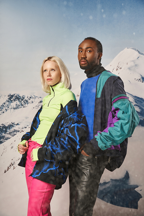 attractive diverse couple in stylish warm outfits with snowy mountain on backdrop, winter concept