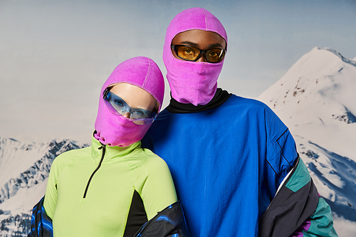 young stylish couple in warm vibrant outfits and pink balaclavas with snowy backdrop, winter concept