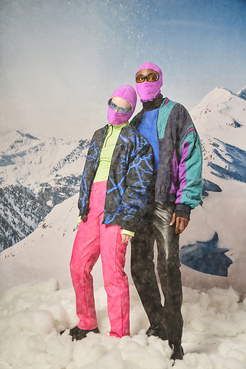 attractive diverse couple posing together in pink balaclavas with mountain backdrop, winter concept
