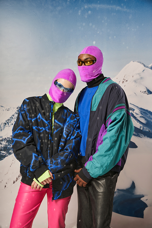 stylish young couple in warm winter outfits and balaclavas posing together on snowy backdrop