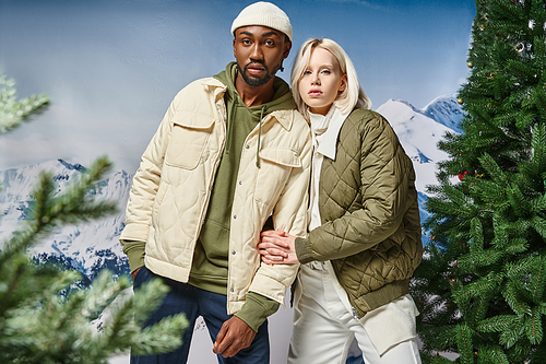 stylish multiracial couple in warm winter clothes posing together on snowy backdrop, fashion concept