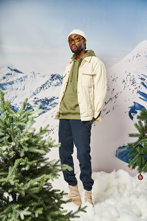fashionable man in stylish warm outfit with beanie hat posing next to pine trees, winter concept