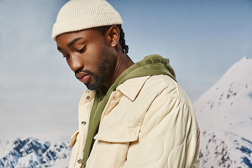 handsome african american man in jacket and beanie looking down with snowy backdrop, winter fashion