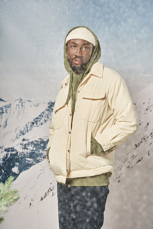 stylish african american man posing with hands in pockets standing under snowfall, winter concept