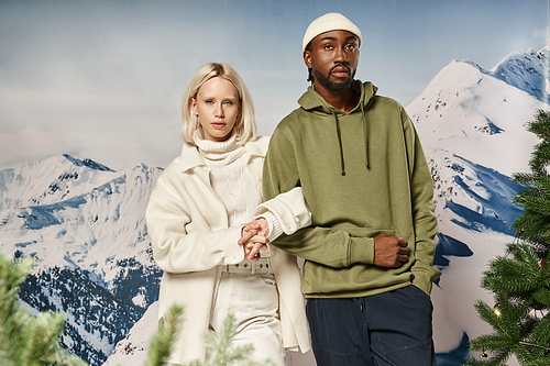 stylish blonde woman standing with african american boyfriend with mountain backdrop, winter fashion