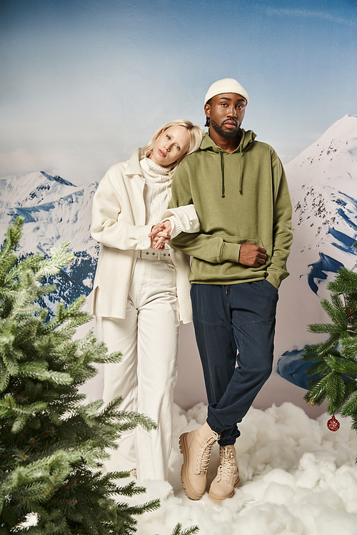full length of stylish interracial couple in winter attire standing together with mountain backdrop