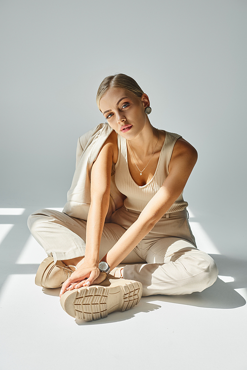 sensual woman in beige clothes and laced boots sitting in sunshine on grey backdrop, full length