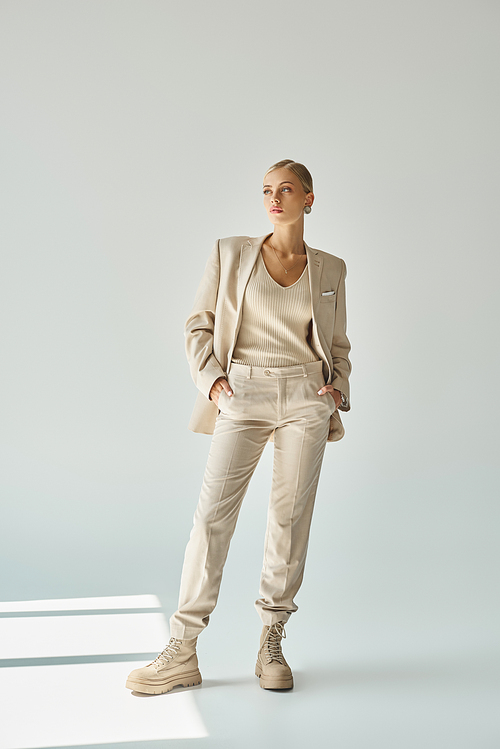 trendy woman in pastel beige suit posing with hands in pockets and looking away on grey, full length