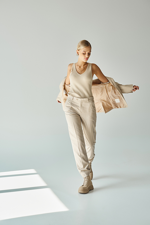 charming woman in total beige outfit posing with stylish blazer on grey backdrop with sunlight