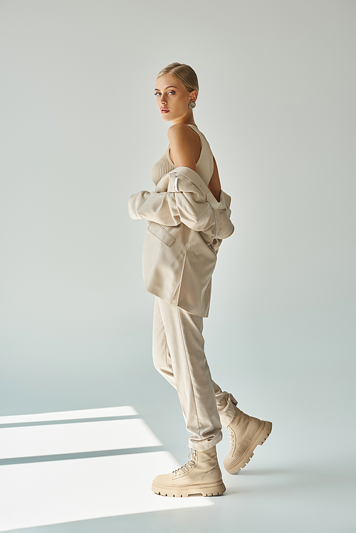 young sensual woman posing in beige suit and laced boots while looking at camera in sunlight on grey