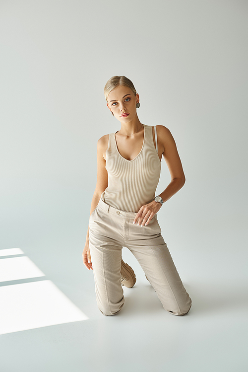 trendy blonde woman in tank top and pants kneeling on grey backdrop with sunlight, urban fashion