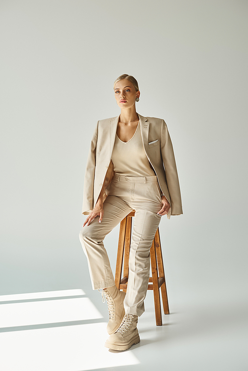 dreamy young woman in pastel beige suit looking away on tall stool in sunlight on grey backdrop