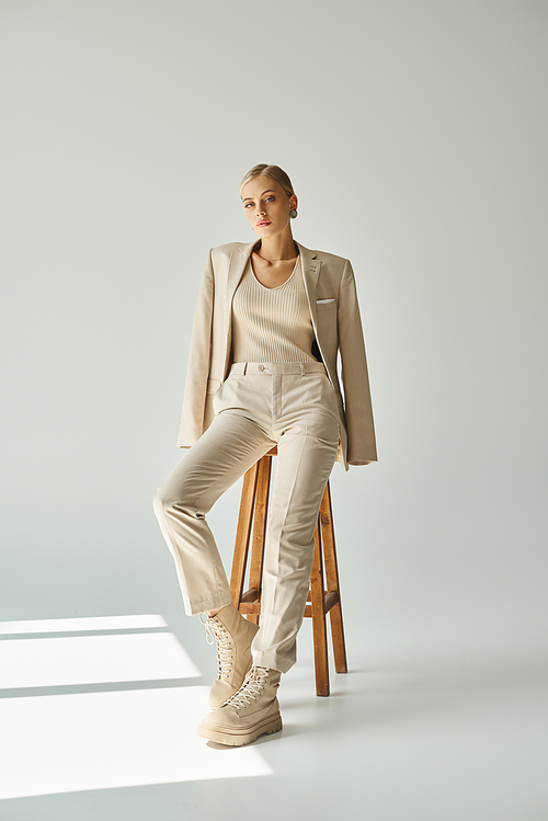 pretty blonde woman in pastel beige suit looking at camera on tall stool in sunshine on grey