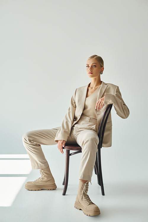 blonde fashion model in pastel beige suit sitting on chair and looking away in sunshine on grey